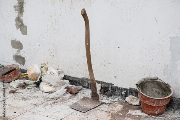 Obraz A hoe in front of a white wall with a dirty floor, around it there is a red bucket, a yellow ladle and a sack as well as bricks and some building debris, photographed from the right angle