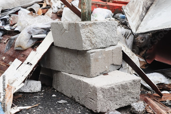 Fototapeta Three white bricks to support green iron rods are stacked on the asphalt, around and behind them are mounds of building remains in the form of plywood, wood and used plastic bags