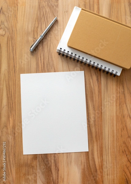 Fototapeta Top-down view of a blank white sheet of paper with spiral notebooks, a mustard-colored journal, and a silver pen on a wooden desk. Ideal for workspace, journaling, or creative mockups.