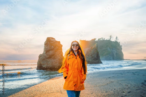 Fototapeta Brunette woman wearing a yellow rain jacket on Ruby Beach posing at sunset. Scenic coastal view with sea stacks, tidepools, and warm twilight colors. Olympic National Park, Washington State.