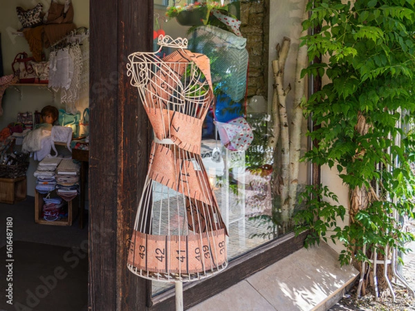 Obraz Elegant tailor's bust with a tape measure waist in front of a charming shop window, flanked by fabrics, accessories, and green vine leaves
