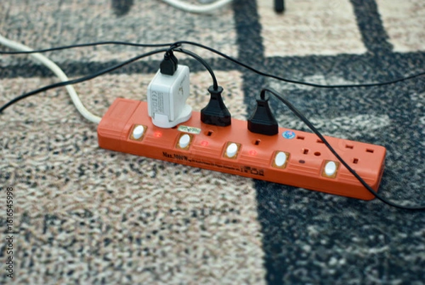 Fototapeta Selangor, Malaysia - February 20, 2025: A multi-outlet power strip, cords snaking across a patterned rug.