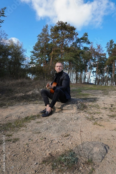 Obraz Portrait of smiling caucasian artist plays wooden acoustic guitar in autumnal park. Young songwriter plays string musical instrument hobby outside in nature fall time. Audio music healing in natural