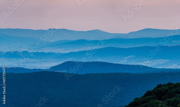 Fototapeta Blue Morning. The mountain ridges of Covasna County, Romania, rise in layered tiers toward the horizon. In the bottom left, a peaceful village lies nestled in the embrace of dawn's first light. 