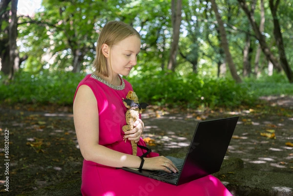 Fototapeta Young woman working on her laptop in a summer park gently cradles her harnessed agama lizard in a tender embrace