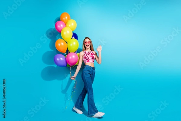 Fototapeta Cheerful young woman holding colorful balloons against a vibrant blue background while walking, expressing fun and happiness