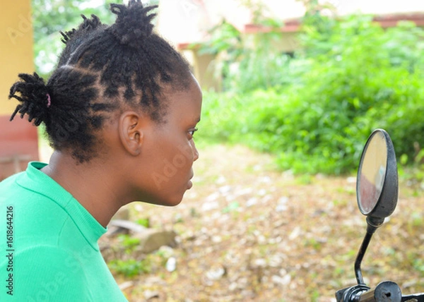 Fototapeta Picture of an African woman in a green top looking into a motorcycle mirror, surrounded by lush greenery in a rural setting.