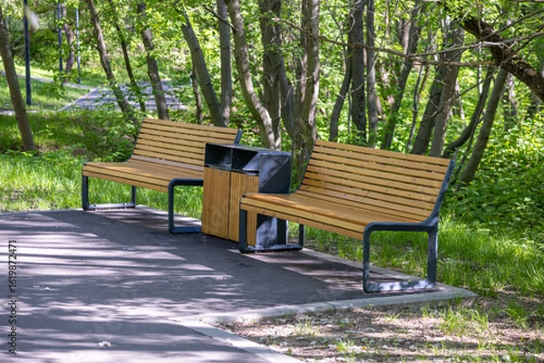 Fototapeta Wooden benches beside a trash bin in a sunny park, surrounded by lush green trees and grass, casting shadows on the paved pathway in a serene outdoor setting