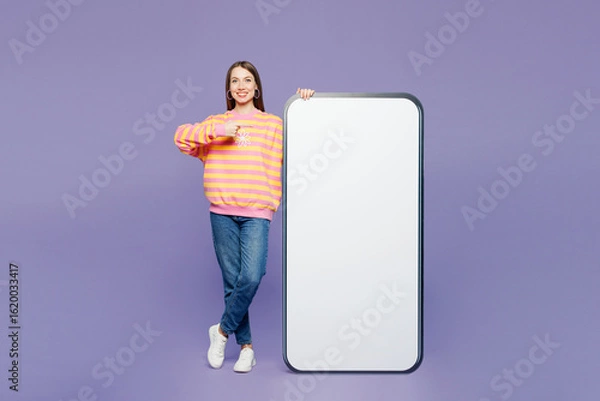 Fototapeta Full body young happy woman wears yellow sweatshirt casual clothes point finger on big huge blank screen mobile cell phone smartphone with area isolated on plain purple background. Lifestyle concept.
