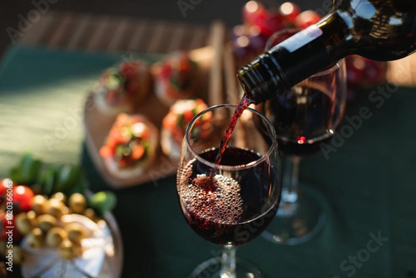 Fototapeta Red wine being poured into a glass on a terrace table with delicious gourmet appetizers in summer