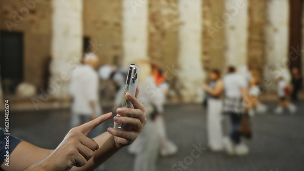 Fototapeta Man using smartphone outdoors on city street capturing moment, with focus on hand and device, people blurred in background for urban lifestyle context.