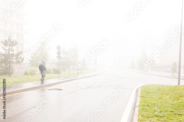 Fototapeta A man walks through a foggy city along an empty road