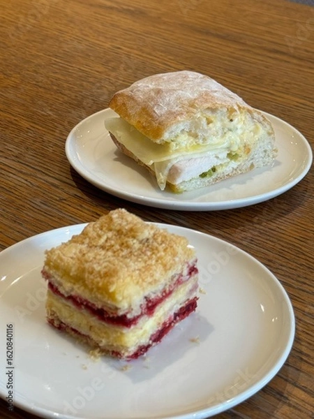 Fototapeta Sandwich and dessert on white plates on wood table at the cafe. Food image, UGC content photo. Breakfast and lunch simple eats at the cafe or home