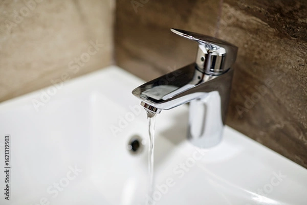 Fototapeta Modern chrome faucet with flowing water in a pristine white sink, surrounded by textured stone wall, showcasing contemporary bathroom design and functionality