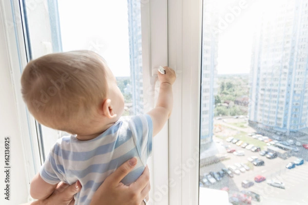 Fototapeta Mother holding baby boy while he is looking outside through window