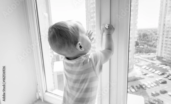 Fototapeta Black and white image of baby in danger. Baby boy pulling handle of window