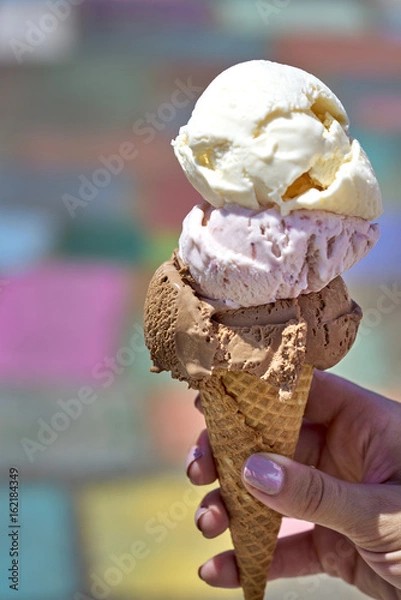 Fototapeta Chocolate, vanilla and berry ice cream scoops in waffle cone held by female hand against colorful background
