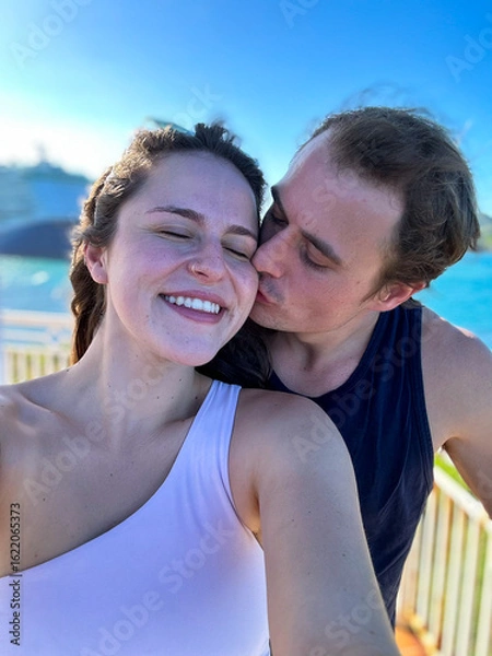 Fototapeta A man kisses his girlfriend on the cheek while she takes a selfie on the ocean coastline.
