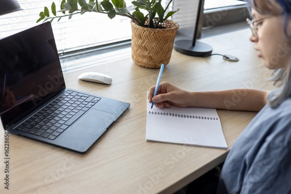 Fototapeta Top view of a student using a laptop and mouse at a desk with notebook and glasses. Modern home study setup in a bright, casual environment.