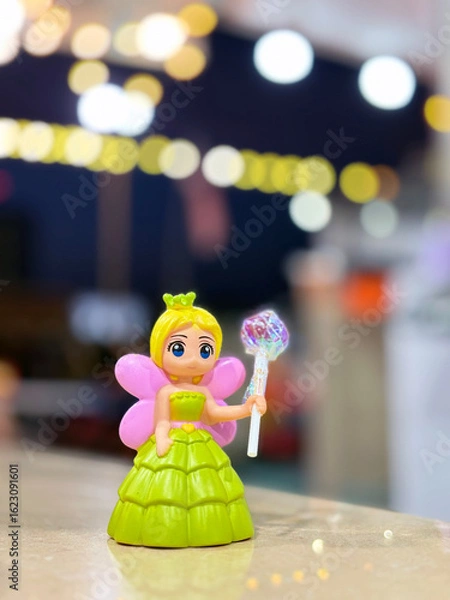 Fototapeta A small, vibrant fairy doll in a green dress with pink wings and a shimmering wand stands on a surface, with blurred, festive bokeh lights in the background.