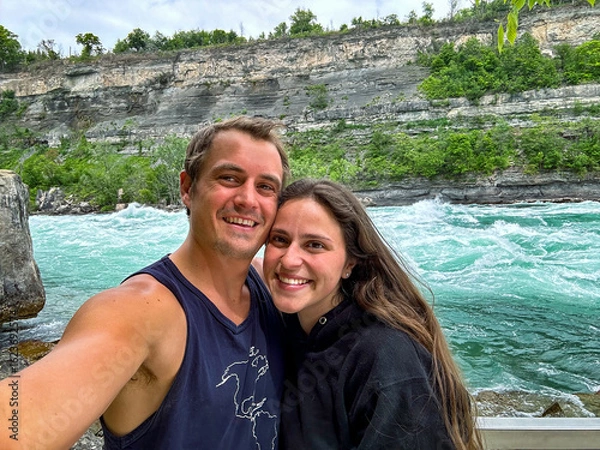 Fototapeta A young couple smiles while taking a selfie with the Niagara rapids and the Escarpment in the background.