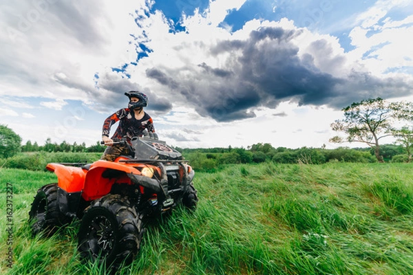 Fototapeta Man on the ATV Quad Bike in a field. Blue sky with clouds.