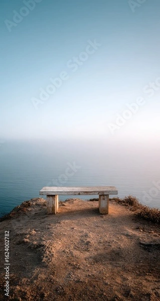 Fototapeta A solitary weathered stone bench sits atop a cliff overlooking a calm, misty ocean under a pale sky