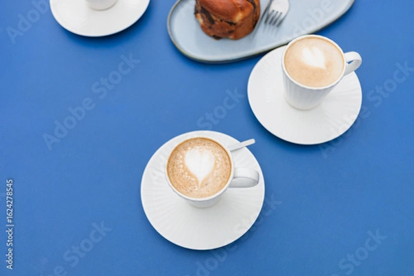Fototapeta Close-up overhead view of cappuccino coffee in beautiful, elegant white cups with pastries placed on a blue table.