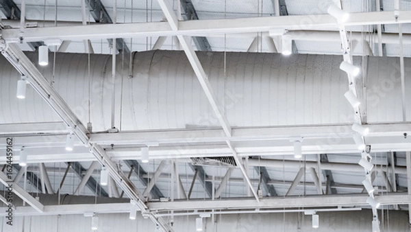 Obraz White LED spotlight lamps hanging on the ceiling frame under steel roof truss structure with air duct pipe system nearby. Lighting design installation in modern warehouse or wide span building