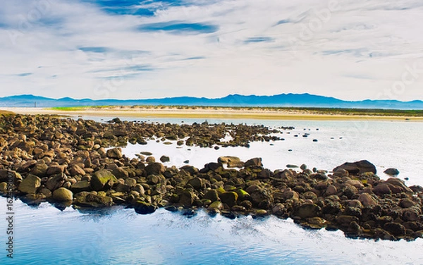Fototapeta Mossy rocks in the foreground frame a serene beach and mountain view on the Cíes Islands, Galicia, Spain. A perfect blend of texture, depth, and natural beauty.