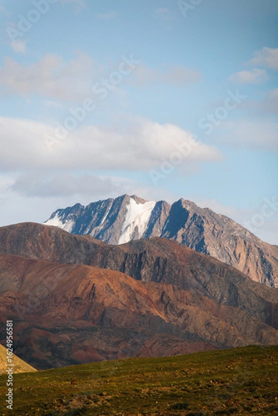 Fototapeta A panoramic view of a rugged, multi-colored mountain range with snowcapped peaks under a clear blue sky with some clouds