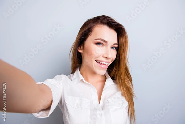 Fototapeta Selfie mania! So hot and pretty! Attractive young lady is making a selfie on the camera, flirty and playful. In casual wear, on pure light background