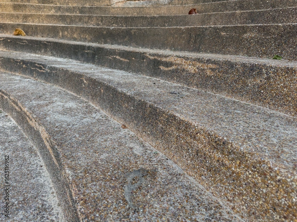 Fototapeta A low-angle view of weathered, curved concrete steps, showing their speckled texture and horizontal lines receding upwards, with a blurred natural background.