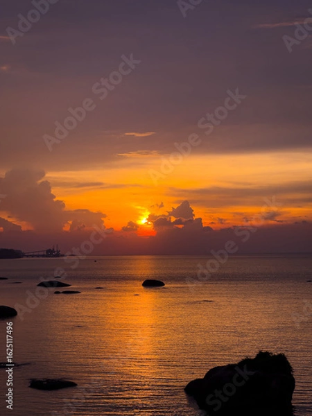 Fototapeta A beautiful orange and purple sunset over the calm sea, with scattered dark rocks in the foreground and a distant city skyline faintly visible on the horizon.