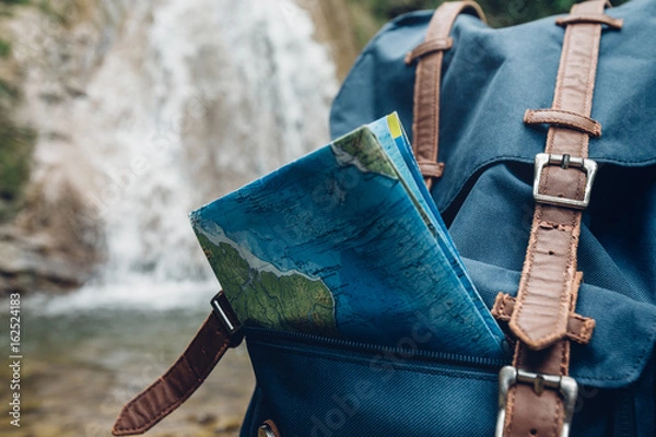Fototapeta Hipster Blue Backpack, And Map Closeup. View From Front Tourist Traveler Bag On Waterfall Background. Exploring Adventure Hiking Concept