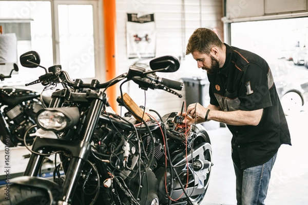 Fototapeta Professional motorcycle mechanic working in bike repair service. Mechanic checking a bike battery level with voltmeter in garage