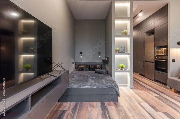 Fototapeta A minimalist bedroom with an integrated kitchen area. The sleeping space is positioned along the wall, next to a work surface with appliances and lighting, creating a cozy atmosphere.