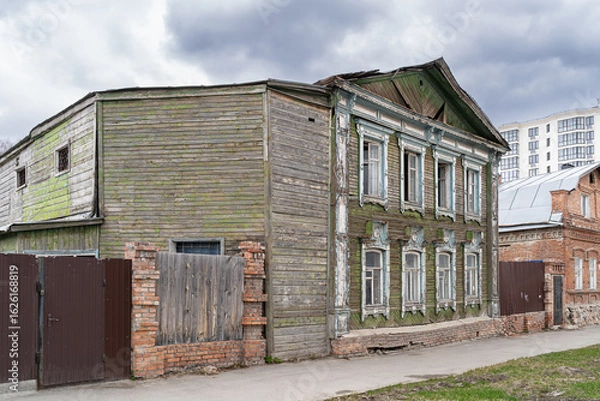 Fototapeta An old wooden house with peeling paint and a modern building in the background create a contrast between the past and the present