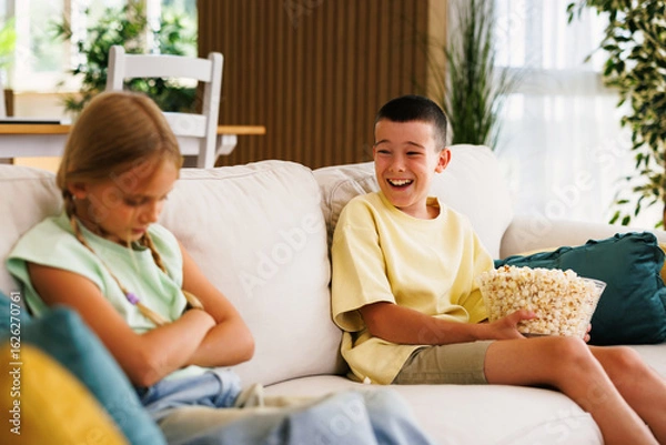 Fototapeta Boy laughing and eating popcorn while sister is angry on sofa