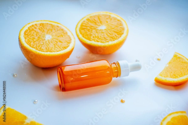Fototapeta Vitamin C skincare concept with a glass bottle of organic facial serum and fresh orange slices arranged on a clean white background. A minimal, natural setup highlighting citrus-based beauty treatment