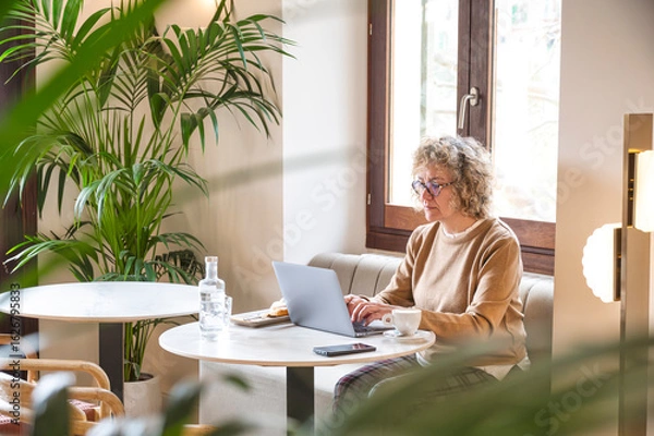 Fototapeta Middle-aged woman working remotely on laptop from cozy café during vacation, enjoying peaceful atmosphere and natural light while balancing work and leisure