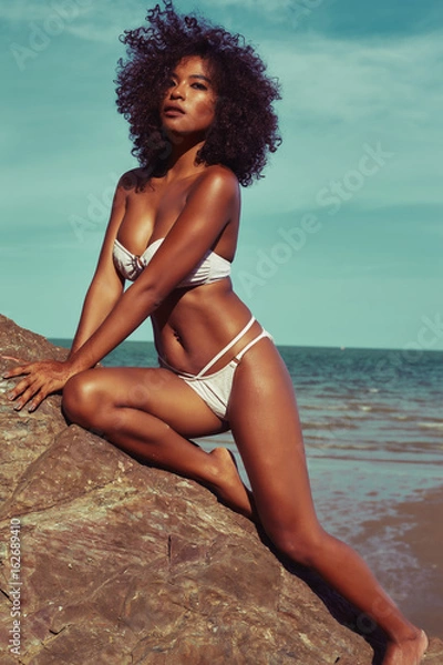 Obraz Egzotyczna modelka z opaloną skórą i kręconymi włosami w bikini na kamienistej plaży