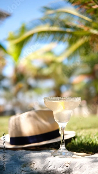 Fototapeta Drinking glass of margarita cocktail decorated lemon slice on table bar, palm trees and blue summer sky on background. Beach bar, summer sea resort. High quality FullHD footage
