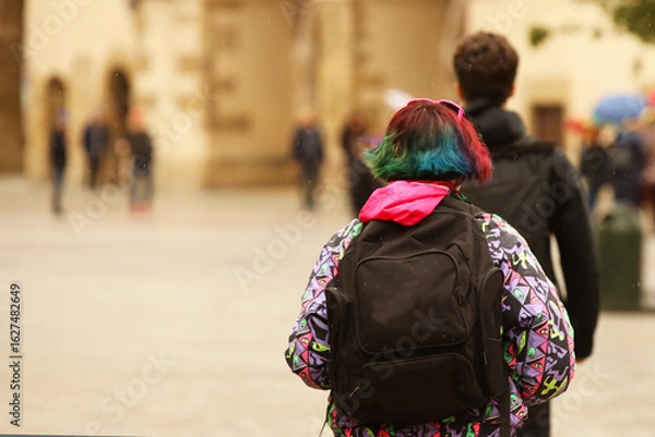 Fototapeta A young girl with multi-colored hair and a trendy haircut walks around the city with a backpack. Teenage protest and the war of generations. Challenge to society. Originality of behavior. Challenging