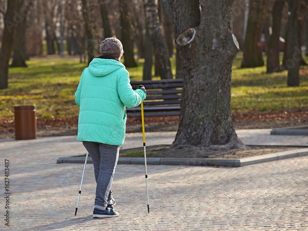 Fototapeta Nordic walking. An elderly woman in urban clothes is walking in the park in the rays of sunlight. Healthy lifestyle. Sporting event. Activity of the elderly. Wellness exercises in the fresh air