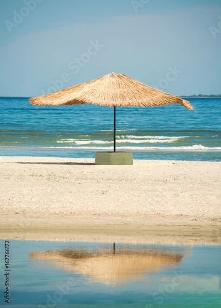 Fototapeta Słomiany parasol przed słońcem na plaży z odbiciem w zatoce morza