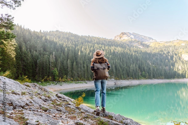 Fototapeta A back view of a female hiker in a hat and with a backpack sitting on a rock overlooking a high mountain lake. Young woman feels freedom and enjoys mountain landscape. Hiking and nature concept.