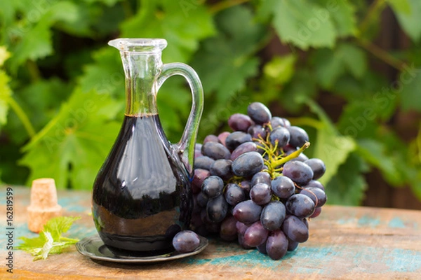 Fototapeta Black old balsamic vinegar in a glass jug with fresh red grapes on green vineyard background