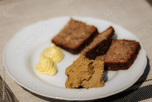 Fototapeta Plate with three slices of dark rye bread, a serving of liver pâté, and two decorative swirls of butter on a white dish atop a beige placemat.