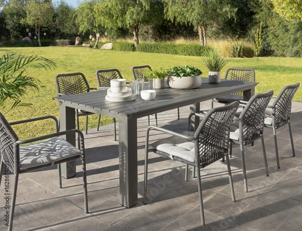 Fototapeta Idyllic outdoor dining set in a verdant garden, a sleek modern slate-gray aluminum table, stylish woven rope chairs with cushions, perfectly placed for a refreshing morning breakfast under the sun.